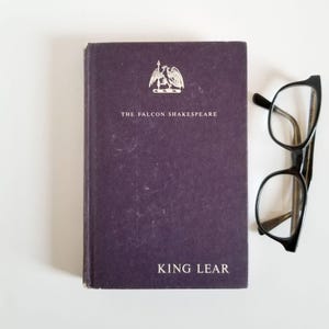 May include: A purple hardback book with the title "King Lear" and the text "The Falcon Shakespeare" on the front cover. A pair of black-framed glasses is lying on a white surface next to the book.