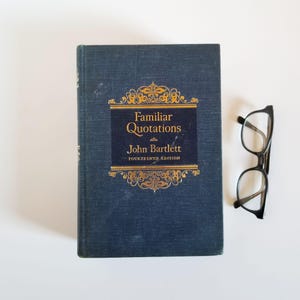 May include: A navy blue hardback book with gold lettering titled "Familiar Quotations" by John Bartlett, Fourteenth Edition. A pair of black-framed spectacles is to the right of the book.