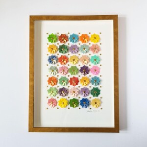A framed artwork featuring a grid of colorful paper flowers, each with a gold center. The flowers are arranged in a random pattern and are set against a white background. The frame is gold. The artist's signature is visible in the bottom right corner of the artwork.
