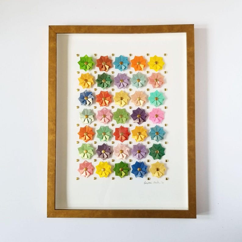 A framed artwork featuring a grid of colorful paper flowers, each with a gold center. The flowers are arranged in a random pattern and are set against a white background. The frame is gold. The artist's signature is visible in the bottom right corner of the artwork.
