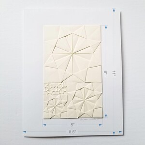 an origami artwork of ivory coloured paper on a white background. The artwork is a geometric design made of folded paper squares. The design is made up of several different shapes, including circles, squares, and triangles. The design is symmetrical and has a radial pattern. The artwork is signed in the bottom right corner. The artwork is shown with dimensions for the collage size as 5 x 8 inches and the background sheet is 8.5 x 11 inches.