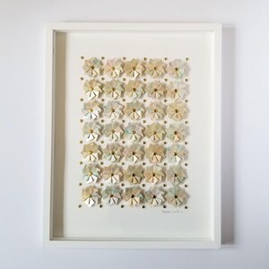 A white framed artwork with a white background and a grid of 35 small origami flowers in shades of beige, pink, and blue. The flowers are arranged in a grid pattern and are accented with gold tone metal pins. The artwork is signed in the bottom right corner.
