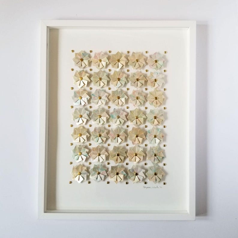A white framed artwork with a white background and a grid of 35 small origami flowers in shades of beige, pink, and blue. The flowers are arranged in a grid pattern and are accented with gold tone metal pins. The artwork is signed in the bottom right corner.