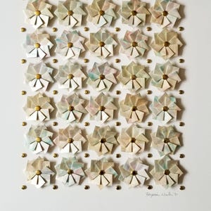 A close up of the bottom portion of the artwork showing small origami flowers mounted on a white background with gold metal pins. The flowers are arranged in a grid pattern. The artwork is signed in the bottom right corner.