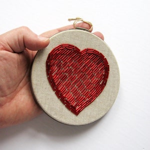 May include: A red beaded heart embroidered on a round piece of beige fabric, framed in a wooden hoop.
