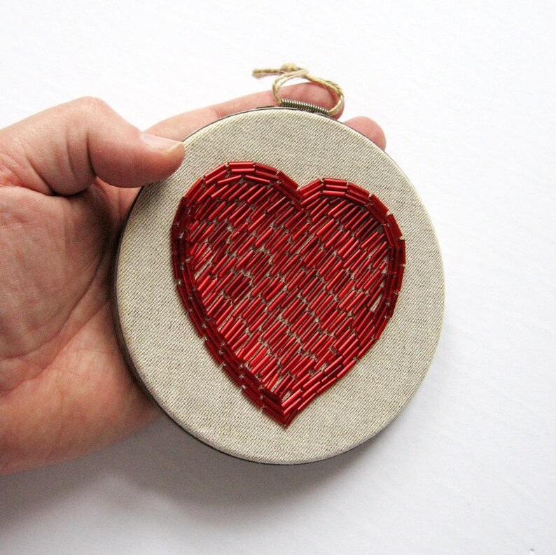May include: A red beaded heart embroidered on a round piece of beige fabric, framed in a wooden hoop.