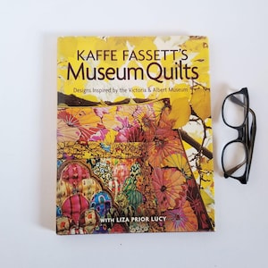 May include: A yellow book titled "Kaffee Fassett's Museum Quilts: Designs Inspired by the Victoria & Albert Museum" with a colourful quilt design on the cover. The book is on a white surface with a pair of black spectacles to the right.
