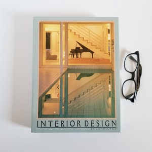 May include: A book titled "Interior Design" by John F. Pile. The cover features a photograph of a modern home with a grand piano and a staircase. The book is on a white surface with a pair of spectacles.