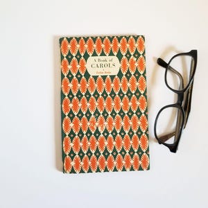 May include: A vintage book titled "A Book of Carols" with a green and orange geometric pattern cover. Black-framed spectacles are placed beside the book on a white surface. The book's cover has a repeating pattern of orange leaf-like shapes.