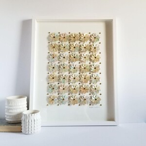 A white framed artwork with a white background and a grid of 35 small origami flowers in shades of beige, pink, and blue. The flowers are arranged in a grid pattern and are accented with gold tone metal pins. The artwork is signed in the bottom right corner.