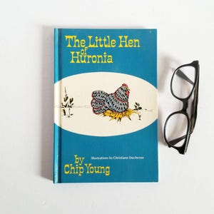 May include: A blue book with the title "The Little Hen of Huronia" by Chip Young. The book cover features an illustration of a gray hen with red wattle and comb sitting on a bed of straw. The book is next to a pair of black eyeglasses.