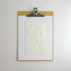 an origami artwork of ivory coloured paper on a white background. The artwork is a geometric design made of folded paper squares. The design is made up of several different shapes, including circles, squares, and triangles. The design is symmetrical and has a radial pattern. The artwork is signed in the bottom right corner. It is shown on a clipboard for size reference.