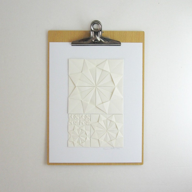 an origami artwork of ivory coloured paper on a white background. The artwork is a geometric design made of folded paper squares. The design is made up of several different shapes, including circles, squares, and triangles. The design is symmetrical and has a radial pattern. The artwork is signed in the bottom right corner. It is shown on a clipboard for size reference.