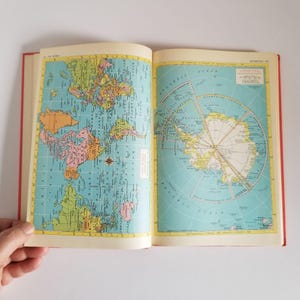 May include: An open book displaying two pages of maps. The left page shows a colorful world map with blue oceans and multicolored continents. The right page features a circular map of Antarctica with yellow landmasses against a light blue ocean.