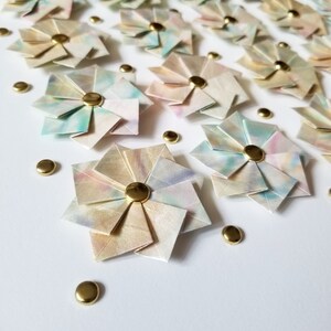 A close up of the bottom left portion of the artwork showing small origami flowers mounted on a white background with gold metal pins.