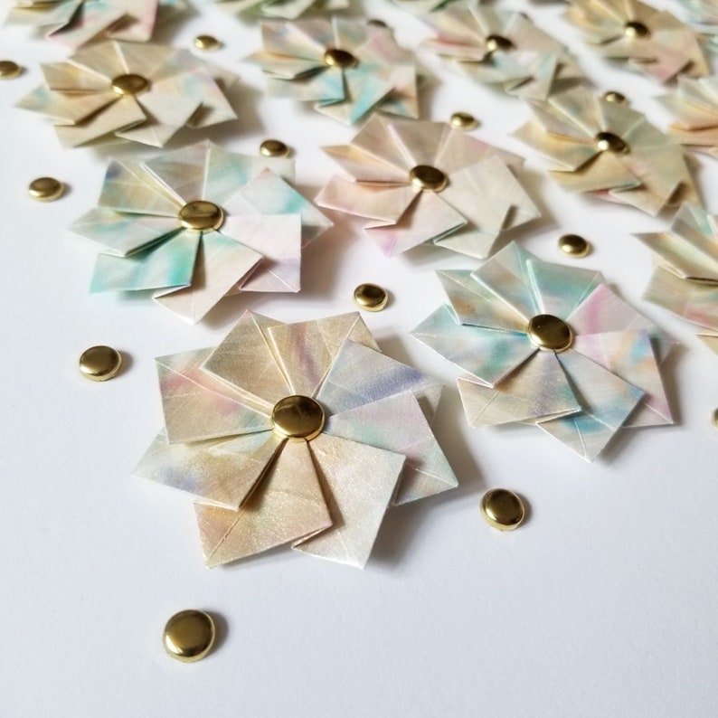 A close up of the bottom left portion of the artwork showing small origami flowers mounted on a white background with gold metal pins.