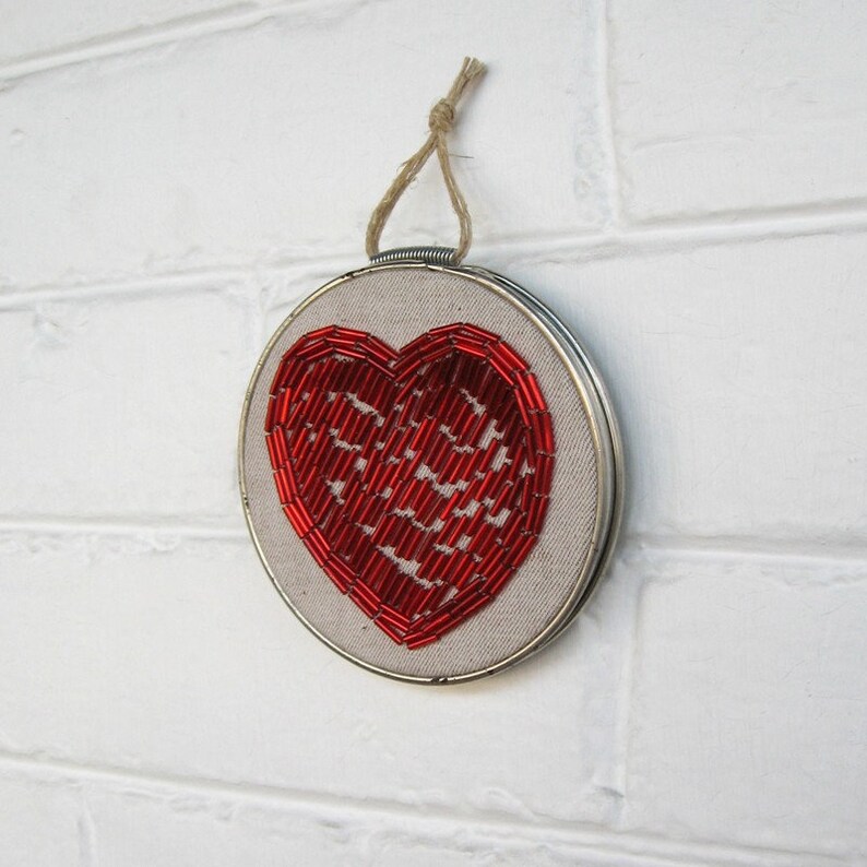 May include: A red beaded heart embroidered on a white canvas, framed in a silver hoop with a twine hanger.