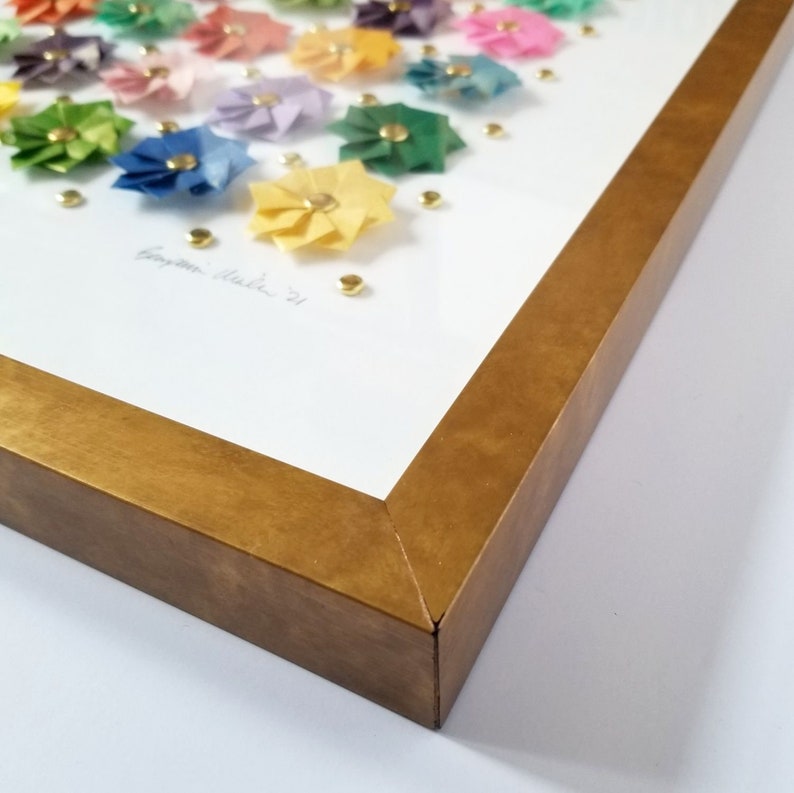 corner detail of a gold framed artwork. The artwork features a collection of colorful paper flowers arranged in a grid pattern. The flowers are made of different color papers, including pink, blue, yellow, and green. The artwork is signed in the bottom left corner.