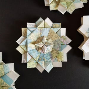 May include: Origami starburst art pieces made from vintage map paper. The central starburst is surrounded by folded paper shapes, showcasing a variety of colors from the maps. The pieces are arranged on a black surface.
