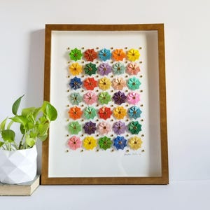 May include: A framed artwork featuring a grid of colorful origami flowers in various hues, including pink, orange, green, and purple. The flowers are mounted on a white background and secured with gold-colored pins. The frame is a warm gold color.