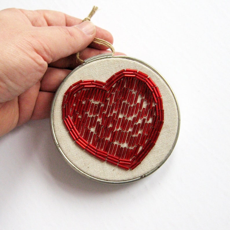 May include: A red beaded heart embroidered on a white canvas, framed in a silver hoop with a brown twine hanger.