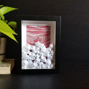 May include: A framed art piece featuring white origami stars cascading from the top. The background has a red and white marbled design with the word "LUCKY!" in white. The frame is black.