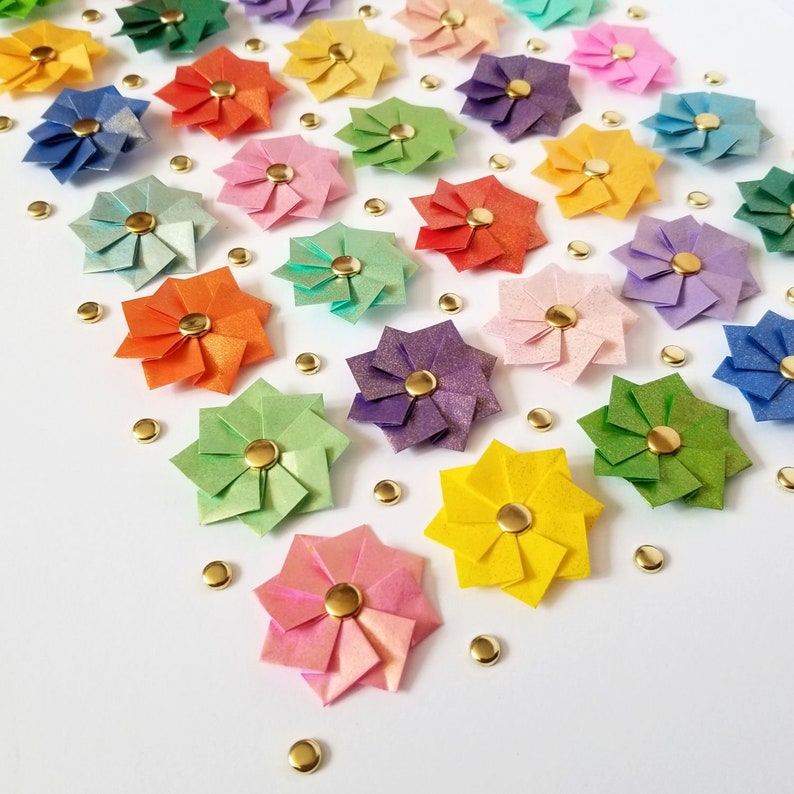 A white surface with origami paper flowers in various colors, including red, orange, yellow, green, blue, purple, and pink. The flowers are arranged in a grid pattern.