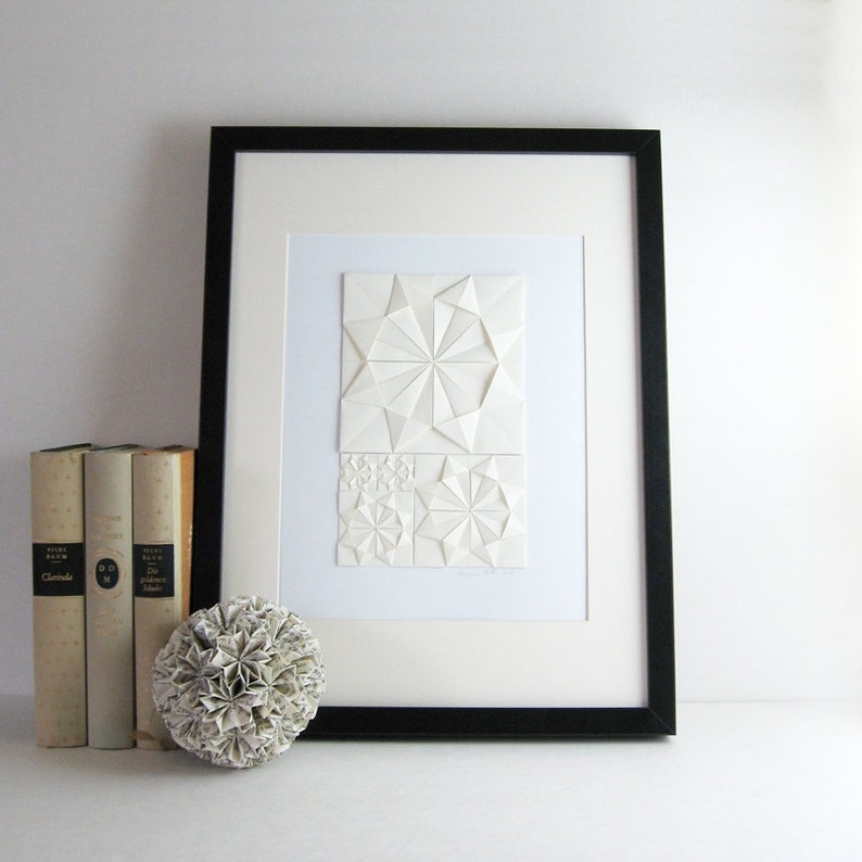 an origami artwork of ivory coloured paper on a white background. The artwork is a geometric design made of folded paper squares. The design is made up of several different shapes, including circles, squares, and triangles. The design is symmetrical and has a radial pattern. The artwork is signed in the bottom right corner. The artwork is shown in a black frame with mat, not included. The art piece is displayed on a white shelf with three books and a white paper ball for size reference.