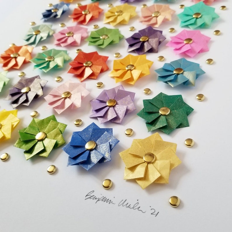 A white background with a rainbow of origami flowers arranged in a grid pattern. The flowers are made of paper and have a gold center. Signed by the artist at the bottom right.