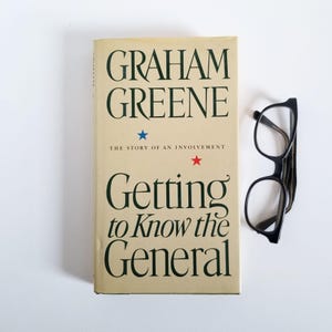 May include: A paperback book titled "Getting to Know the General" by Graham Greene. The book cover is beige with green text and two stars, one blue and one red. The book is lying on a white surface next to a pair of black spectacles.