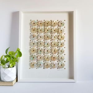 A white framed artwork with a white background and a grid of 35 small origami flowers in shades of beige, pink, and blue. The flowers are arranged in a grid pattern and are accented with gold tone metal pins. The artwork is signed in the bottom right corner.