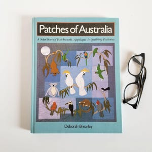 May include: A book titled "Patches of Australia" with a teal cover featuring patchwork designs of Australian animals and birds. Black-framed glasses rest beside the book. The book's author is Deborah Brearley.