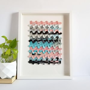 May include: A framed wall art piece featuring a geometric paper weaving pattern in shades of pink, light blue, black, and white. The artwork is composed of interlocking folded paper strips arranged in a repeating chevron-like grid inside a white frame.