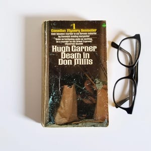 Peut inclure: Une couverture de livre de poche avec un fond brun et une photo de sacs en papier brun. Le titre du livre est "Death in Don Mills" par Hugh Garner. Le livre est un best-seller du mystère canadien.