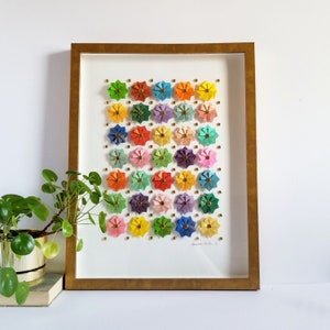 A framed artwork featuring a colorful arrangement of paper flowers in a grid pattern. The flowers are in shades of pink, purple, blue, green, yellow, and orange. The frame is gold and the artwork is signed bottom right.