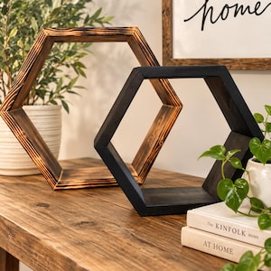 May include: Two hexagonal wooden wall shelves. One shelf features a natural wood grain with charred, dark edges, while the other is finished in a solid matte black. Both shelves have an open, geometric frame design with six sides.