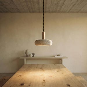 May include: A pendant light fixture featuring a smooth, rounded, light-colored stone or concrete shade suspended by a dark, twisted fabric cord. The fixture has a cylindrical wooden connector at the top where the cord meets the shade.