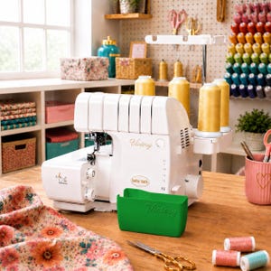 Könnte beinhalten: Eine weiße Baby Lock Victory Overlock-Nähmaschine mit vier gelben Garnrollen, einem grünen Kunststoff-Abfallbehälter und dem aufgedruckten Text Victory und Jet-Air Threading, platziert auf einer Holzoberfläche mit verschiedenen Nähutensilien.