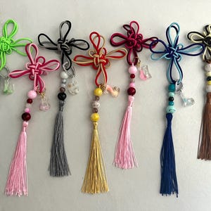 Puede incluir: Siete colgantes decorativos con nudos de cuerda complejos en verde neón, rosa, negro, rojo, magenta, azul y crema. Cada colgante incluye una borla, cuentas variadas de diferentes colores y texturas, y un pequeño dije de resina con forma de letra.