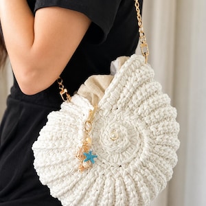 Peut inclure: Un sac à main en crochet blanc en forme de coquillage, doté d'une bandoulière à chaîne dorée, d'une perle centrale et d'une breloque décorative avec une petite étoile de mer bleue et des perles. Une doublure en tissu beige est visible.
