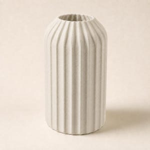 Könnte beinhalten: Eine zylindrische Vase mit matter, cremeweißer Oberfläche und einer texturierten, vertikal gerippten Außenseite. Das Gefäß verfügt über eine schmale, kreisförmige Öffnung an der Oberseite und einen geraden, gleichmäßigen Körper.