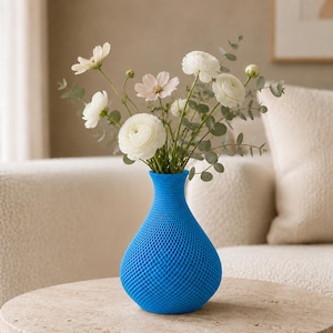 Könnte beinhalten: Eine leuchtend blaue Vase mit einer strukturierten, kreuzschraffierten geometrischen Oberfläche. Die Vase hat einen schmalen Hals und einen abgerundeten Boden und ist mit einem Strauß aus weißen und blassrosa Blumen sowie grünen Eukalyptusblättern gefüllt.