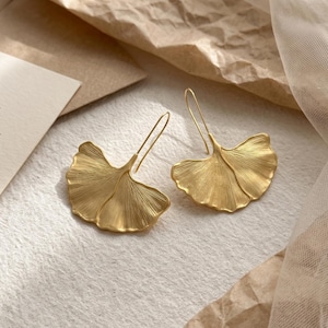 May include: A pair of gold-toned drop earrings shaped like ginkgo leaves. Each earring features a textured, fan-like surface with fine, radiating lines and a slender, curved wire hook for attachment, showcasing a metallic finish.