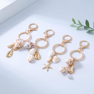 Puede incluir: Cuatro llaveros de metal en tono dorado, cada uno con una anilla circular y un cierre de mosquetón. Los llaveros están decorados con varias conchas de colores naturales, pequeñas estrellas de mar blancas y pequeñas perlas blancas.