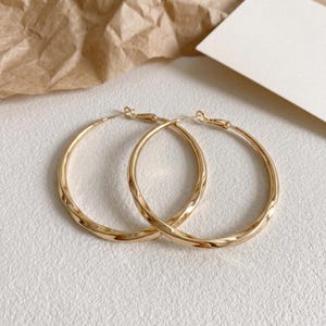 May include: A pair of gold-toned hoop earrings featuring a twisted, textured metal design. The circular earrings have a polished, reflective finish and include a standard hinged post closure mechanism at the top of each hoop.