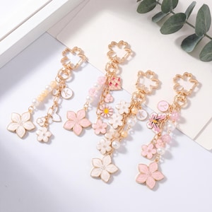 Puede incluir: Cuatro llaveros de metal dorado con motivos de flores de cerezo, cuentas tipo perla colgantes y pequeños dijes. Cada llavero incluye una anilla en forma de flor, colgantes de flores esmaltadas en tonos rosa y blanco, y un dije circular con la letra K.
