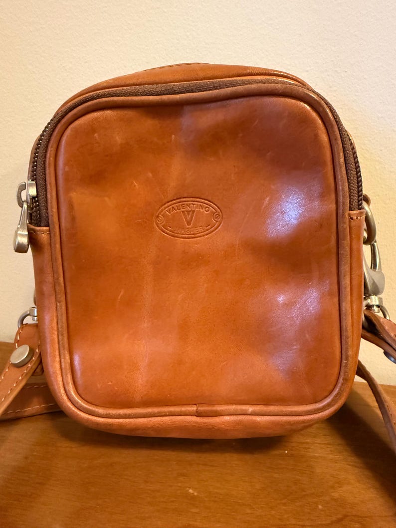 Może przedstawiać: Jasnobrązowa sk&oacute;rzana torebka na ramię o prostokątnym kształcie z zaokrąglonymi rogami. Z przodu znajduje się wytłoczone owalne logo z napisem VALENTINO MASTER. Posiada ciemnobrązowy zamek błyskawiczny, srebrne okucia oraz dopasowany pasek.