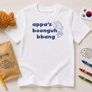 May include: A white short-sleeved t-shirt featuring the dark blue text "appa's boonguh bbang" and two small fish illustrations. The shirt is surrounded by a plush fish toy, a paper bag with Korean text and a fish graphic, crayons, and a flag sticker.