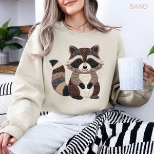May include: A sand-colored crewneck sweatshirt featuring a central graphic of a raccoon designed with a patchwork quilt aesthetic. The raccoon illustration uses various shades of brown, tan, and black with dotted and floral patterns.
