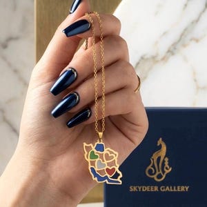 May include: Gold-colored necklace with a pendant shaped like a map of Iran, featuring colorful heart-shaped designs. The necklace is held by a hand with long, dark blue nails. A navy blue box with gold text is in the background.